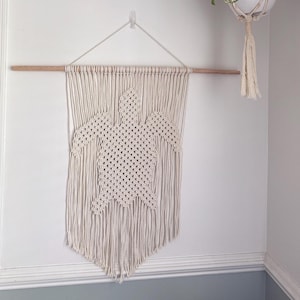 May include: Cream-colored macrame wall hanging featuring a turtle design. The wall art is suspended from a wooden dowel and hangs against a white wall. A white hanging planter with green plants is also visible.