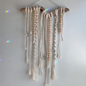 Op de afbeelding: Een macramé wandhanger met een basis van drijfhout, bestaande uit witte katoenen koorden, houten kralen en kristallen accenten.