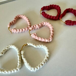 May include: Three pairs of heart-shaped hoop earrings made of braided cord. The earrings are in pink, white, and red.