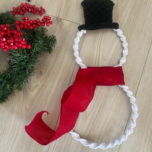 May include: A white crocheted snowman wreath with a black crocheted hat and a red ribbon.