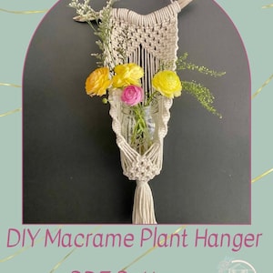 May include: DIY macrame plant hanger PDF pattern. A white macrame plant hanger with a wooden dowel at the top and a tassel at the bottom. A glass jar is inside the hanger, holding yellow and pink flowers.