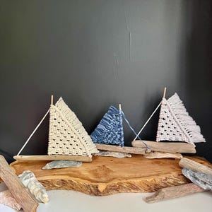 May include: Three handcrafted macrame sailboats with driftwood bases. The sails are cream, blue, and cream, and the boats are arranged on a wooden slab. Decorative driftwood and stones surround the boats, creating a nautical theme.