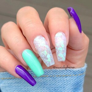 May include: Close-up of a hand with long, light blue, green, and purple nails with glitter accents. The nails are long and coffin shaped. The hand is holding a piece of denim fabric. The text "@manisbydesiree" is visible on the fabric.