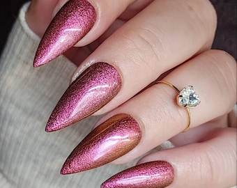 May include: Close-up of a hand with long, pointed, burgundy and gold glitter nails. The nails are painted with a gradient effect, with the burgundy color at the tips and the gold color at the base. The hand is wearing a gold ring with a heart-shaped diamond.