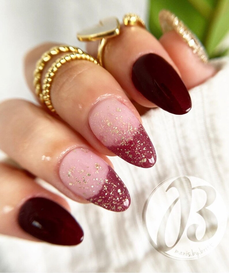 May include: Close-up of a hand with almond-shaped nails painted in a gradient design. The nails are painted with a light pink base and a burgundy tip with gold glitter. The nails are accented with a gold glitter design.