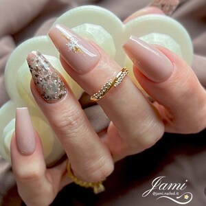 May include: Close-up of a hand with long, nude-colored coffin-shaped nails. One nail features a glittery, silver and pink design, while another has a delicate gold snowflake design. A gold ring with small diamonds is worn on the ring finger.