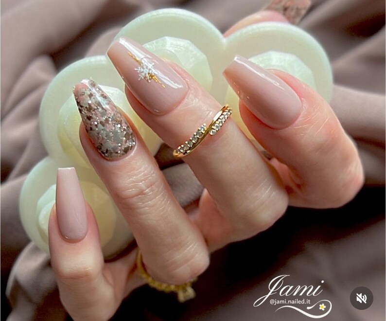 May include: Close-up of a hand with long, nude-colored coffin-shaped nails. One nail features a glittery, silver and pink design, while another has a delicate gold snowflake design. A gold ring with small diamonds is worn on the ring finger.