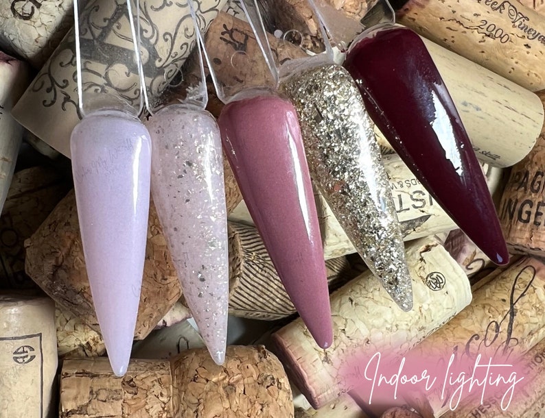 May include: Five different nail polish colors displayed on a cork background. The colors are a light purple, a light pink with glitter, a darker pink, a gold glitter, and a dark red.