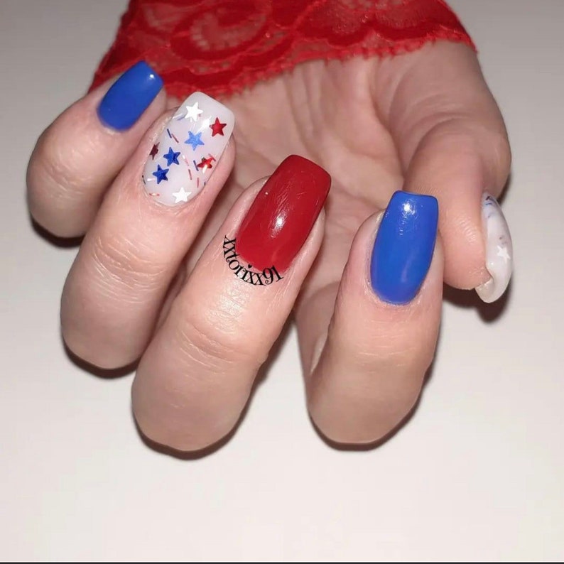 May include: A hand with a manicure featuring red, white, and blue nail polish. One nail is red, two are blue, and one is white with red and blue star-shaped confetti. The index finger nail is white with a marble design. The text "xtorix91" is on the red nail.