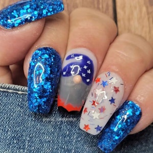 May include: A hand with a manicure featuring blue glitter and patriotic nail art. One nail has a gnome design with a blue hat and stars. Another nail has red, white, and blue star-shaped glitter.