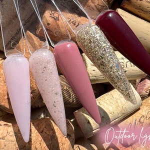 May include: Four nail polish bottles with different colors and finishes. The bottles are arranged in a row on a background of wine corks. The colors are a light pink, a light pink with glitter, a light pink with a shimmer, and a dark red.