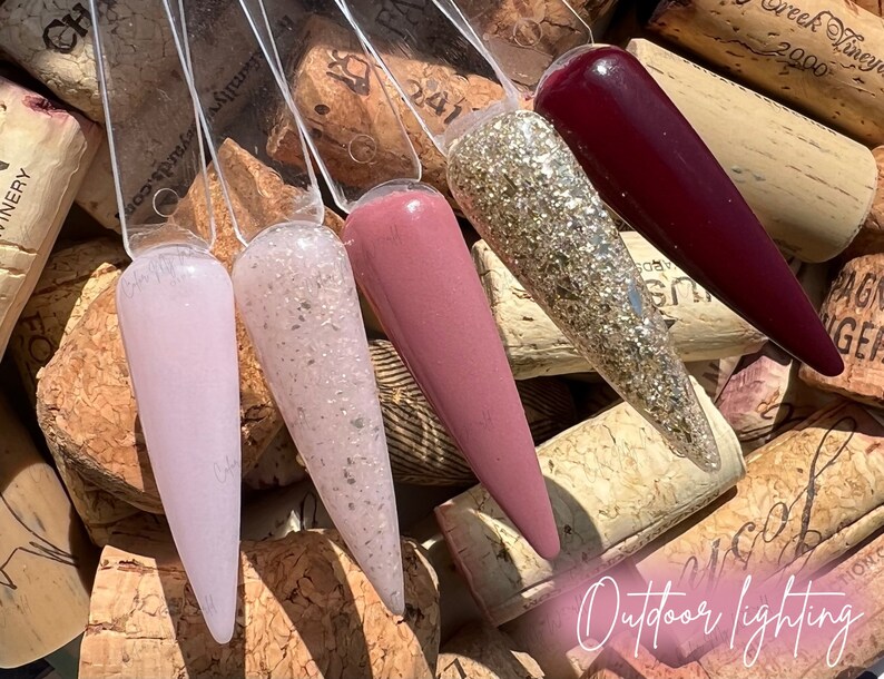 May include: Four nail polish bottles with different colors and finishes. The bottles are arranged in a row on a background of wine corks. The colors are a light pink, a light pink with glitter, a light pink with a shimmer, and a dark red.