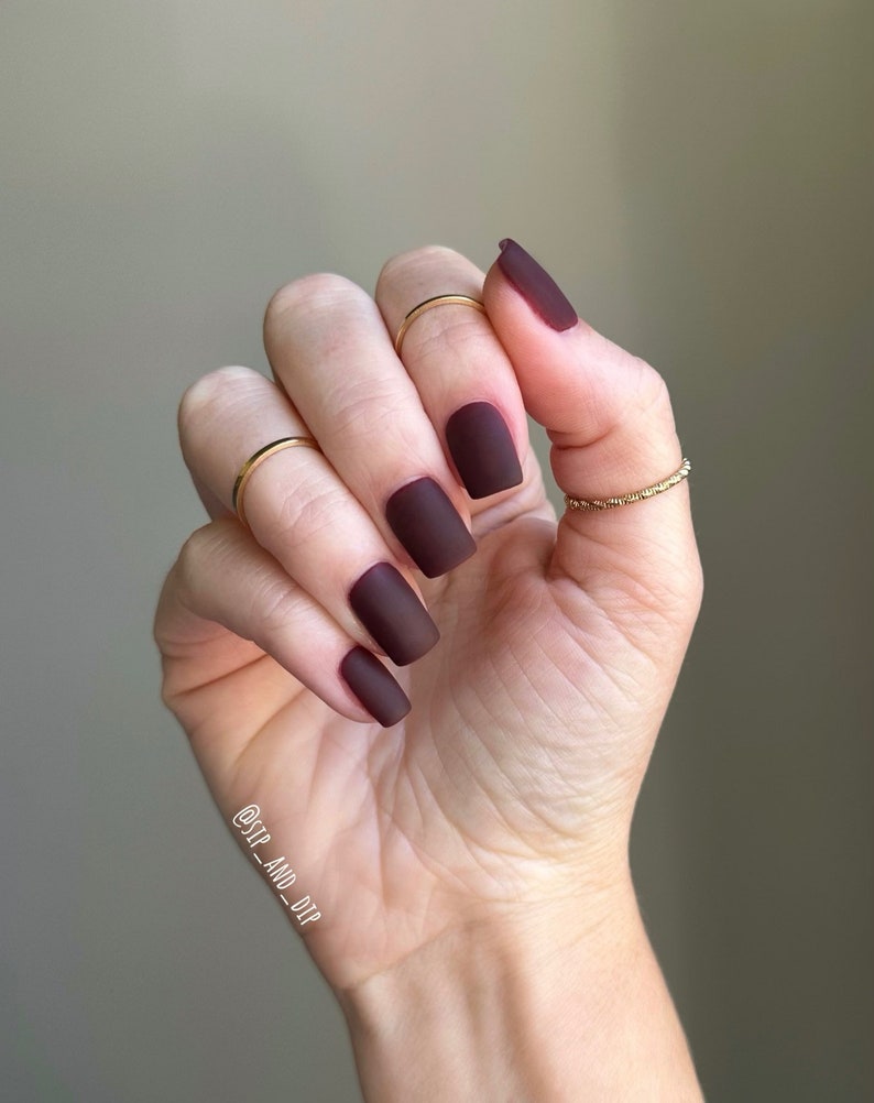 May include: Close-up of a hand with short, square-shaped nails painted a deep burgundy red. The nails have a matte finish.
