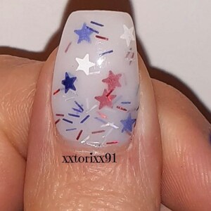May include: Close-up of a fingernail with a white base and festive nail art. The design features red, white, and blue star-shaped glitter and small, colorful confetti. The text "xxtorixx91" is printed on the nail.