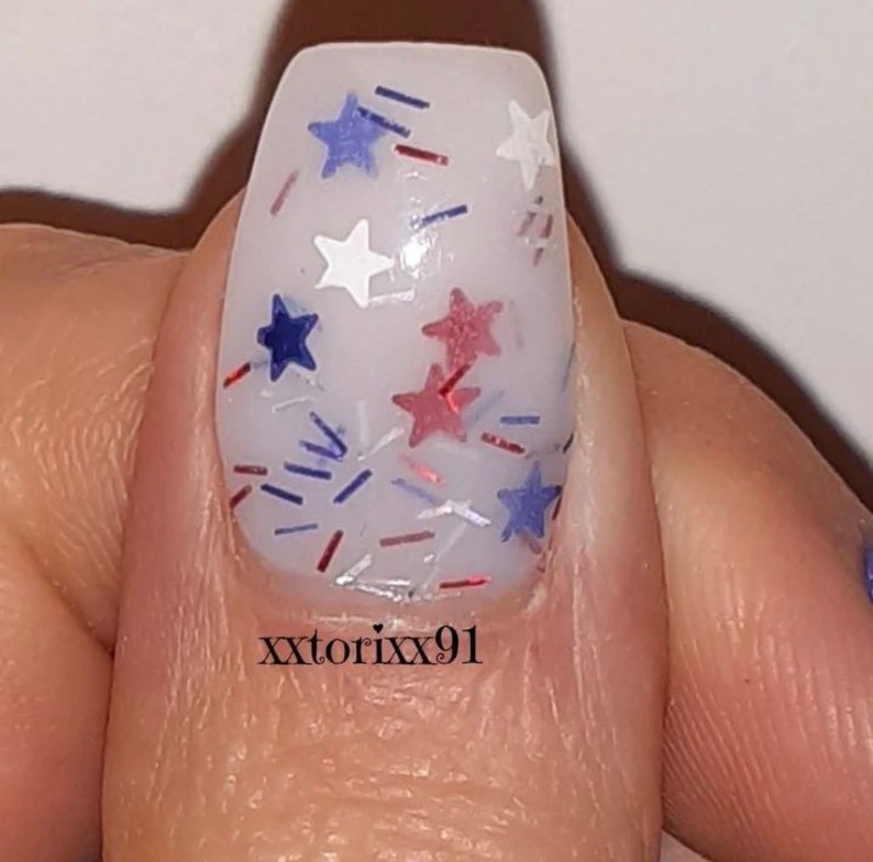 May include: Close-up of a fingernail with a white base and festive nail art. The design features red, white, and blue star-shaped glitter and small, colorful confetti. The text "xxtorixx91" is printed on the nail.