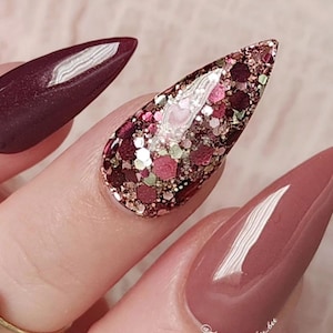 May include: Close-up of a hand with long, stiletto-shaped nails. The nails are painted in a deep burgundy shade and adorned with glitter. The glitter is a mix of gold, pink, and red, creating a festive and glamorous look. The nails are polished to a high shine, giving them a luxurious feel.