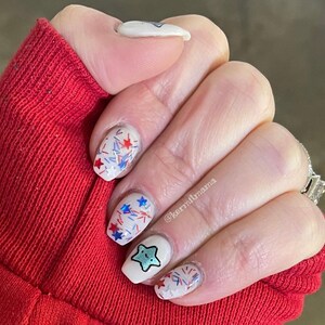 May include: A hand with a red sleeve displays a manicure with white nail polish. The nails are decorated with red, white, and blue star and sprinkle designs, and a cartoon star character. The text "kermitmama" is visible.