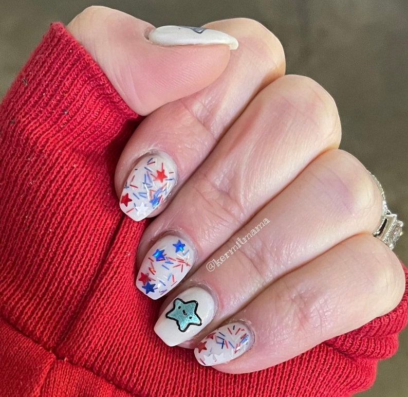May include: A hand with a red sleeve displays a manicure with white nail polish. The nails are decorated with red, white, and blue star and sprinkle designs, and a cartoon star character. The text "kermitmama" is visible.