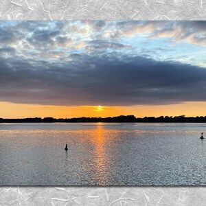 May include: A scenic sunset over a calm body of water with a dark, cloudy sky and a bright orange sun setting behind a line of trees in the distance.