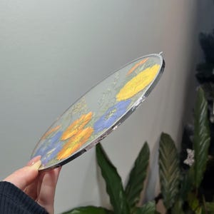 May include: A circular glass wall hanging featuring pressed yellow and blue flowers encased between two clear glass panes, secured with a thin silver-toned metal frame and a small metal loop at the top for hanging purposes.