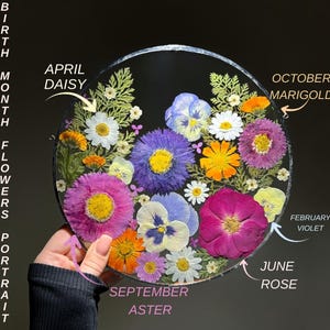May include: A round floral portrait featuring pressed flowers, including daisies, violets, and roses. The text indicates the flowers represent birth months, with labels like "April Daisy" and "October Marigold". The flowers are arranged on a clear, circular base.