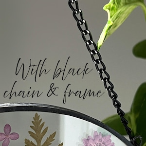 May include: A circular glass hanging ornament featuring pressed purple flowers and green foliage encased within a thin black metal frame. The piece is suspended by a black metal chain and includes the text With black chain & frame.