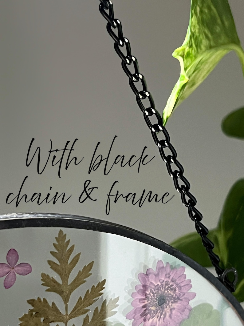 May include: A circular glass hanging ornament featuring pressed purple flowers and green foliage encased within a thin black metal frame. The piece is suspended by a black metal chain and includes the text With black chain & frame.