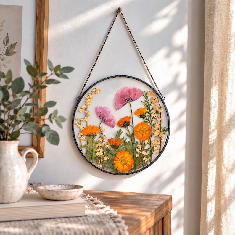May include: A circular wall hanging featuring pressed flowers encased in glass with a dark metal frame. The arrangement includes vibrant orange marigolds, pink poppy petals, and delicate yellow sprigs of greenery suspended by a thin metal chain.