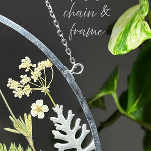 May include: A circular wall hanging featuring pressed dried flowers and greenery encased in a silver frame with a silver chain. The text "With silver chain & frame" is visible. The arrangement includes white flowers and green fern-like leaves.