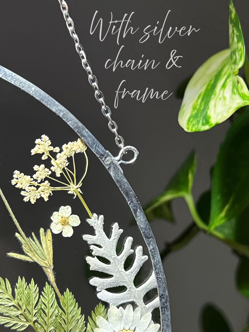 May include: A circular wall hanging featuring pressed dried flowers and greenery encased in a silver frame with a silver chain. The text "With silver chain & frame" is visible. The arrangement includes white flowers and green fern-like leaves.