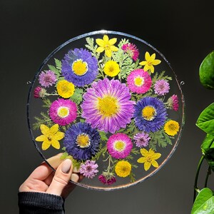 Peut inclure: Une œuvre d'art en résine ronde et transparente présentant un arrangement vibrant de fleurs pressées. Les fleurs sont dans des tons de violet, jaune, rose et bleu, avec du feuillage vert. La pièce est tenue par une main, mettant en valeur le design floral délicat.