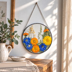 May include: A circular wall hanging featuring pressed flowers in shades of blue, yellow, and orange, encased between two clear glass panes with a thin black metal frame. The piece is suspended by a delicate gold-toned metal chain from a wall hook.