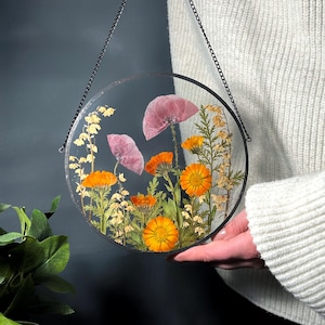 May include: A circular clear resin wall hanging featuring pressed flowers including bright orange calendula, pale pink poppies, and delicate yellow sprigs. The piece is framed in a thin black metal border and suspended by a black metal chain.