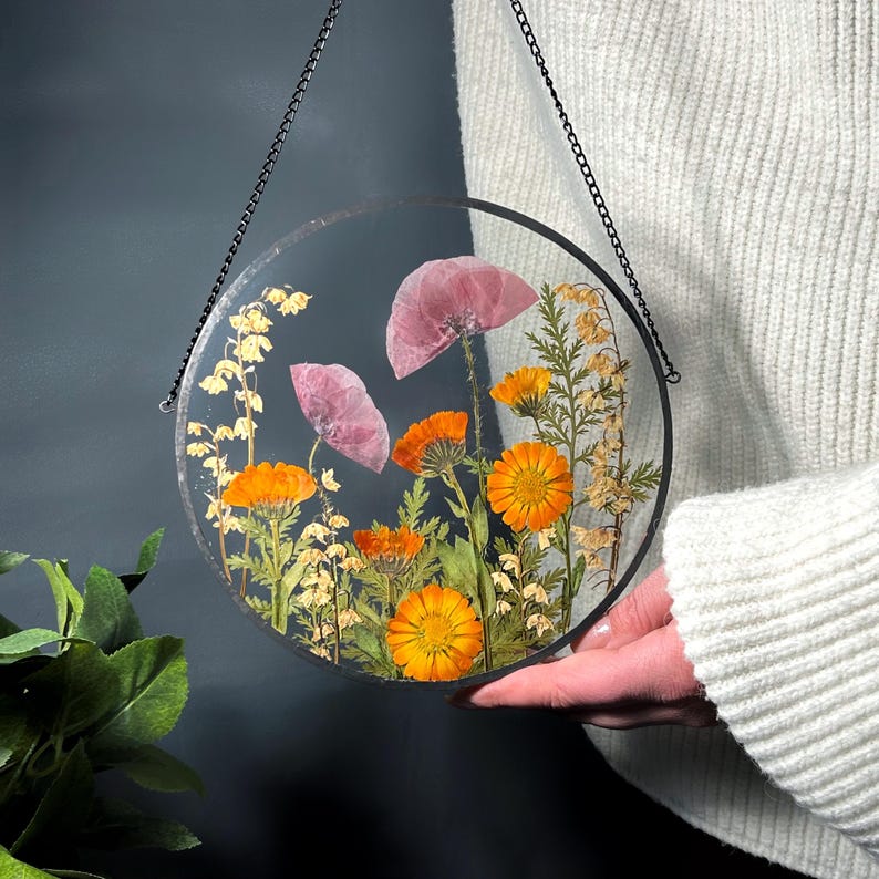 May include: A circular clear resin wall hanging featuring pressed flowers including bright orange calendula, pale pink poppies, and delicate yellow sprigs. The piece is framed in a thin black metal border and suspended by a black metal chain.