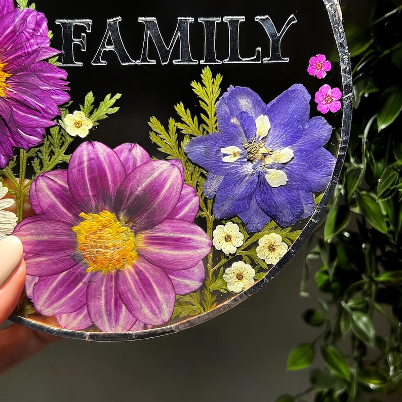 May include: A circular resin plaque featuring the word FAMILY in silver lettering. The design is embedded with pressed purple dahlias, a blue delphinium, small white flowers, tiny pink blossoms, and green fern leaves, all set against a black background.