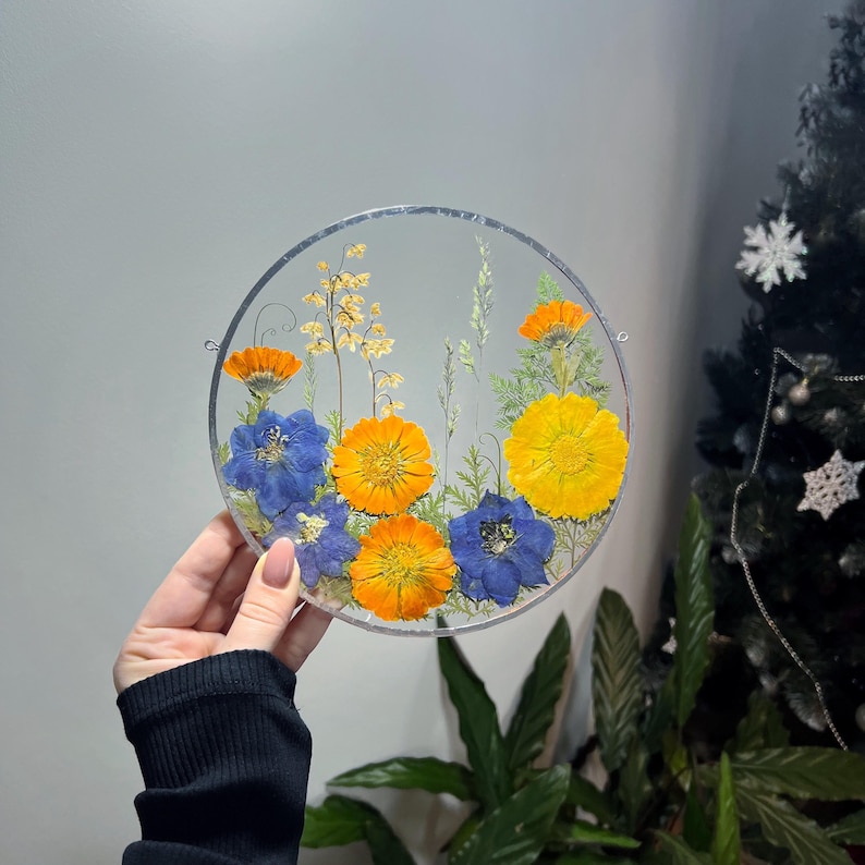 May include: A circular glass wall hanging featuring pressed flowers in vibrant shades of orange, yellow, and blue. The arrangement includes various botanical elements, such as small yellow blossoms, green ferns, and larger orange and blue petals.