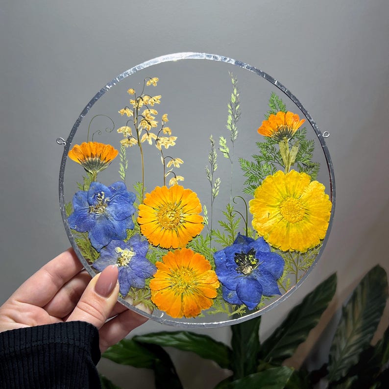 May include: A circular glass wall hanging featuring pressed dried flowers in vibrant shades of yellow, orange, and blue, arranged with green foliage. The arrangement is encased within a thin, silver-toned metal frame with small loops on the sides.