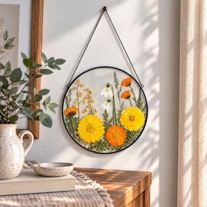 May include: A circular wall hanging featuring pressed yellow and orange flowers with white snowdrop blossoms encased between two clear glass panes within a thin black metal frame, suspended by a matching black metal chain from a small wall hook.