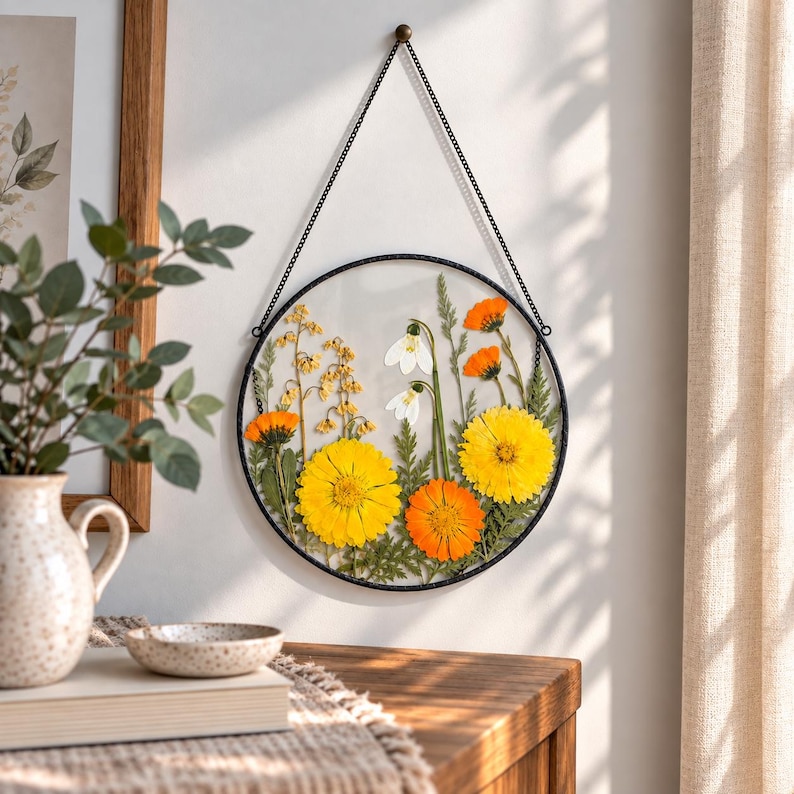 May include: A circular wall hanging featuring pressed yellow and orange flowers with white snowdrop blossoms encased between two clear glass panes within a thin black metal frame, suspended by a matching black metal chain from a small wall hook.