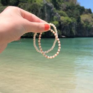 May include: Two white pearl bracelets with orange accents, held in a hand against a backdrop of a tropical beach and lush green foliage.