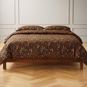 May include: A bed featuring a duvet cover and two matching pillowcases with a brown and black leopard print pattern. The bedding is draped over a simple wooden bed frame with four legs, creating a cohesive look for the sleeping area.