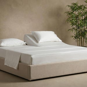 May include: A low-profile platform bed frame upholstered in beige fabric, featuring a split mattress configuration. One side lies flat with a white pillow, while the other side is elevated at an angle, topped with a white pillow and white sheets.