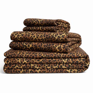 May include: A stack of folded fabric sheets featuring a repeating leopard print pattern in shades of brown, tan, and black. The textile is neatly layered, showing the consistent animal-inspired design across all visible surfaces of the folded items.