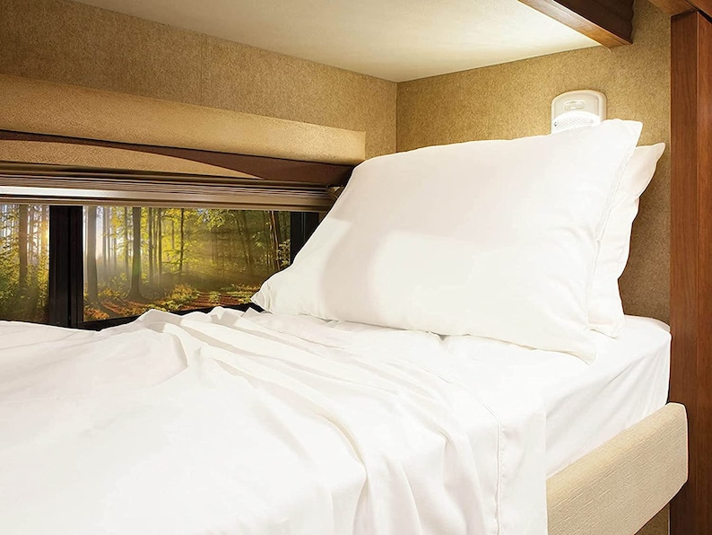May include: A white bed with a white pillow and a white blanket. The bed is in a camper van with a window behind it showing a forest.