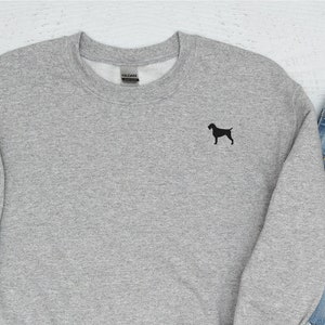 Könnte beinhalten: Ein grauer Pullover mit einer kleinen schwarzen Silhouette eines Hundes auf der Vorderseite. Der Pullover liegt flach auf einer weißen Oberfläche mit einem Lineal, einem Apfel, einer Brille, einem Notizbuch, Jeans und Schuhen drumherum.