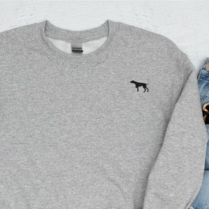 以下が含まれることがあります： 胸に小さな黒い犬のシルエットが刺繍されたヘザーグレーのスウェットシャツ。長袖のクルーネックトップは白い袖口が付いています。画像には、ノート、リンゴ、メガネ、定規、ジーンズ、靴、シュシュが含まれています。