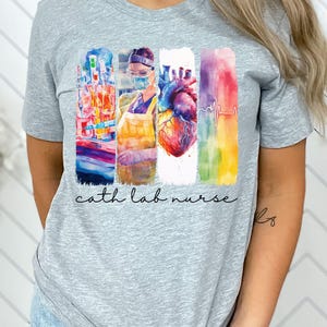 Puede incluir: Camiseta gris jaspeado con un gráfico colorido. El gráfico presenta rayas verticales con ilustraciones médicas y el texto "cath lab nurse". La camiseta está hecha de un material suave.