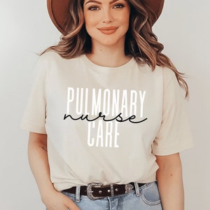 Pode incluir: Uma mulher vestindo uma camiseta bege com o texto "PULMONARY nurse CARE" em preto e branco. Ela está usando um chapéu marrom e jeans azul.