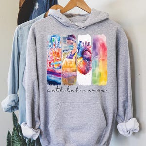 Puede incluir: Una sudadera con capucha gris jaspeado con un gráfico colorido que presenta imágenes médicas y el texto "cath lab nurse". El diseño incluye una enfermera, un corazón y otros elementos médicos. La sudadera tiene capucha y bolsillo delantero.