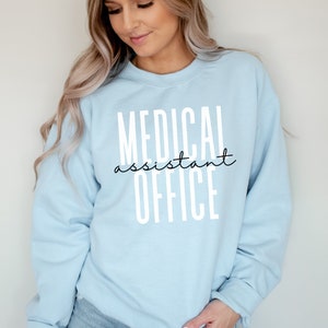 Op de afbeelding: Een lichtblauw sweatshirt met de tekst "MEDICAL assistant OFFICE" in wit gedrukt.